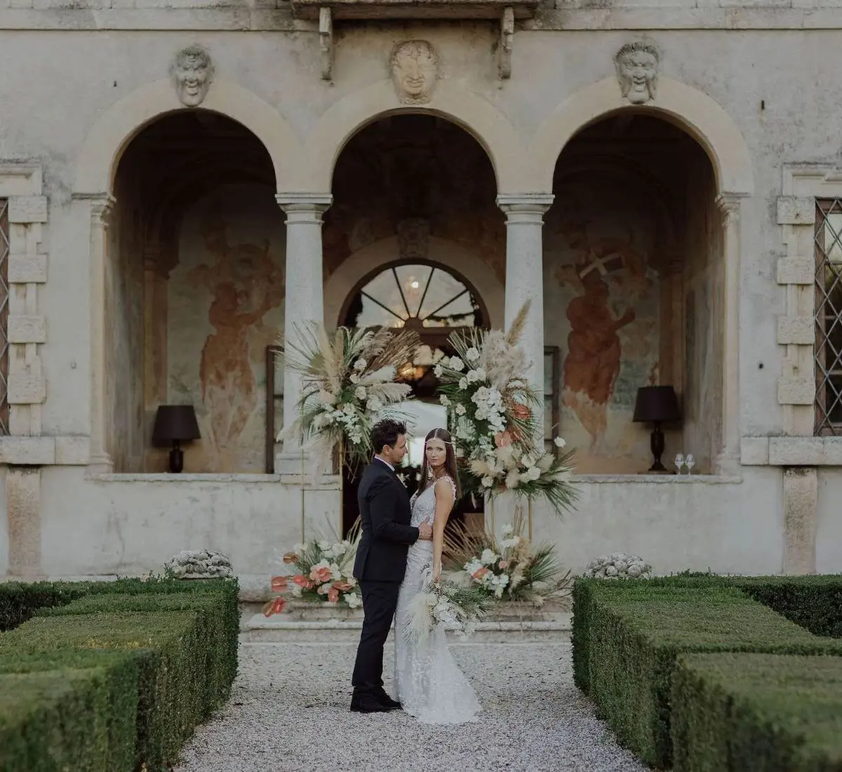 A bride and groom before a frescoed Palladian villa loggia, framed by a pampas-grass installation