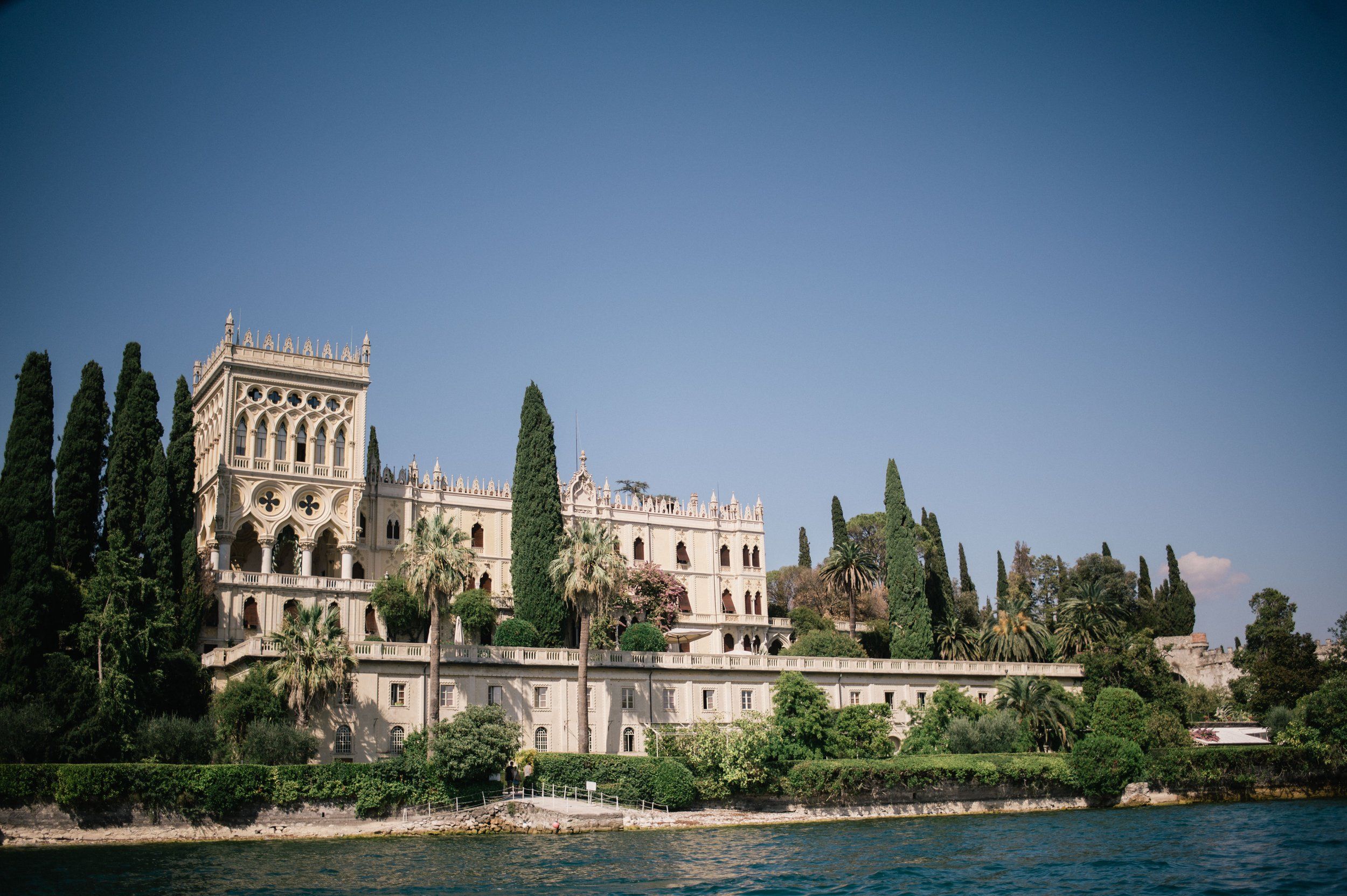 Isola del Garda's neo-Gothic villa seen from the water, cypress and palm trees along the shoreline