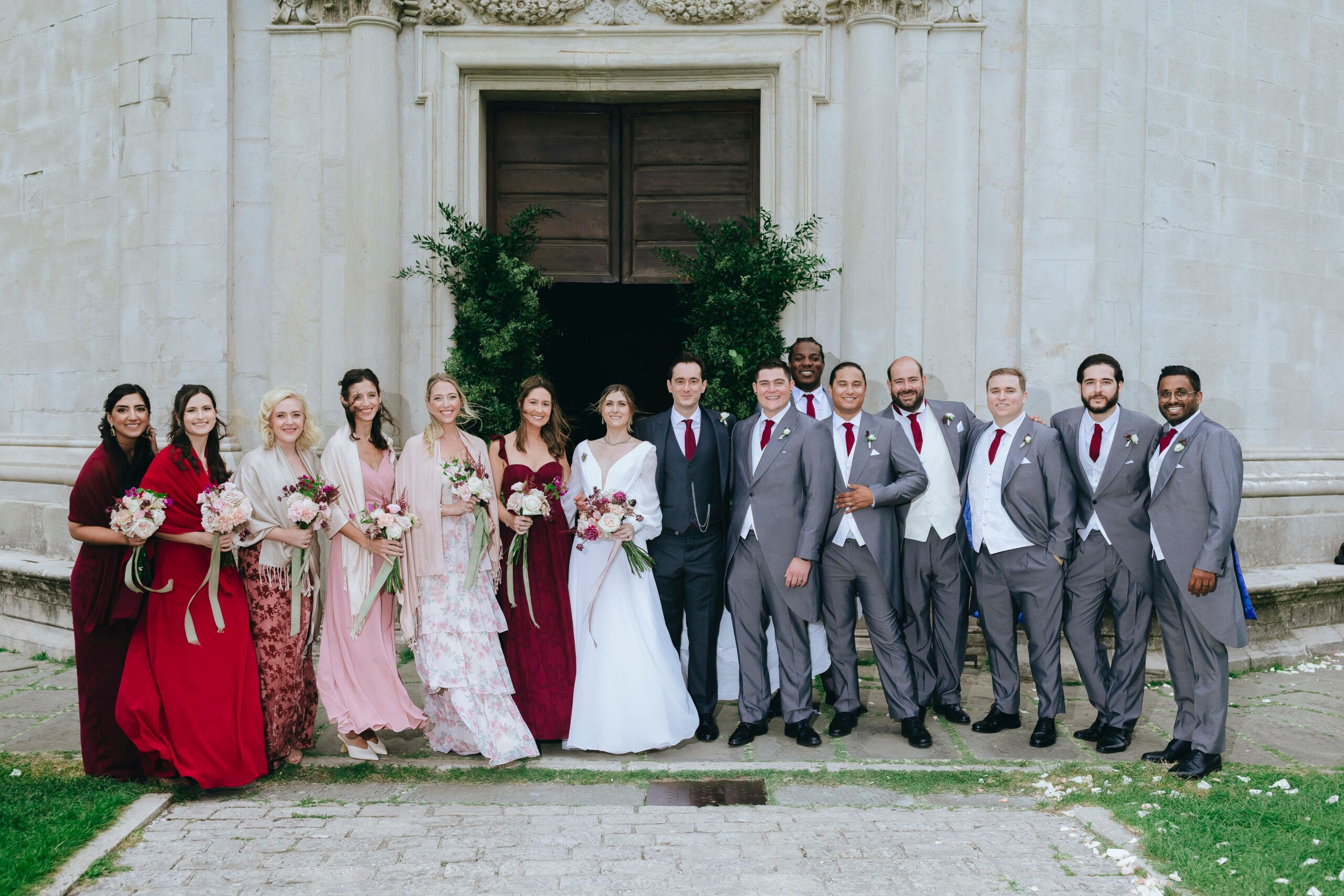 The whole wedding party across a pale church front, burgundy and grey