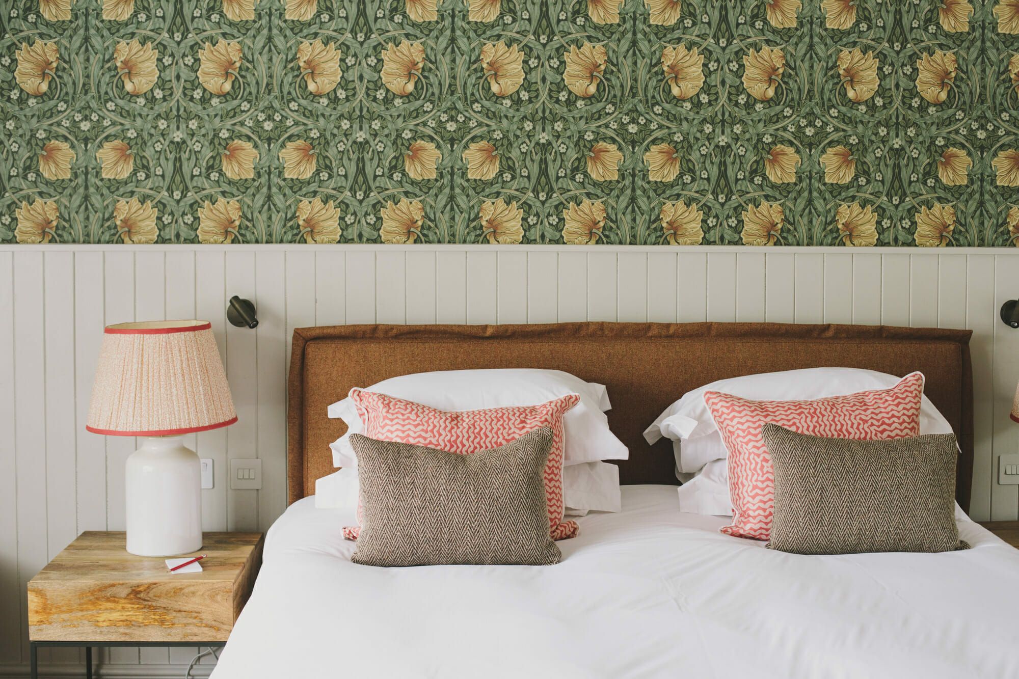 A bedroom in green Morris wallpaper, the bed layered with coral and linen cushions