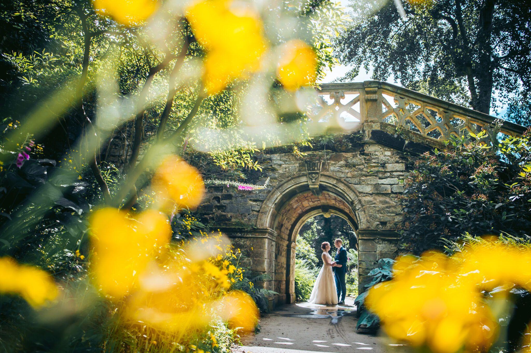 A couple kissing beneath a stone garden archway, framed by yellow flowers