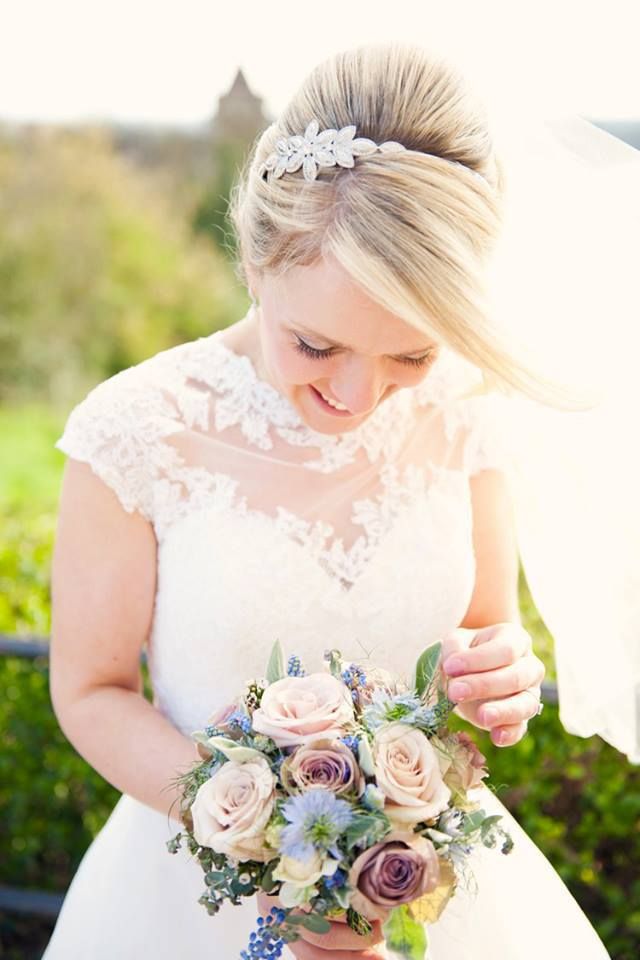 A bride in a lace cap-sleeve gown, holding a dusky pink and lilac bouquet