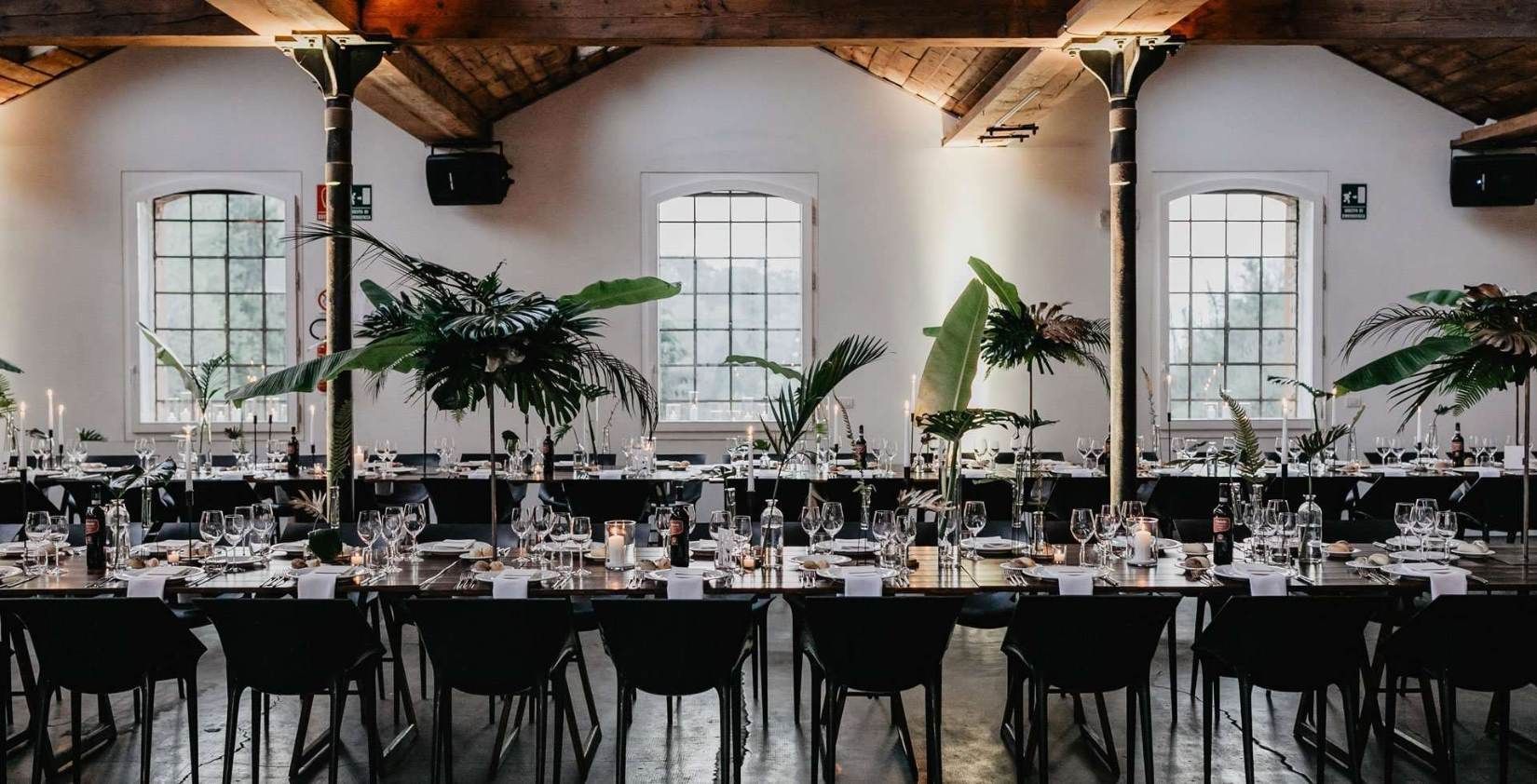 An industrial-chic reception hall: exposed timber beams, black chairs, tall banana-leaf centrepieces