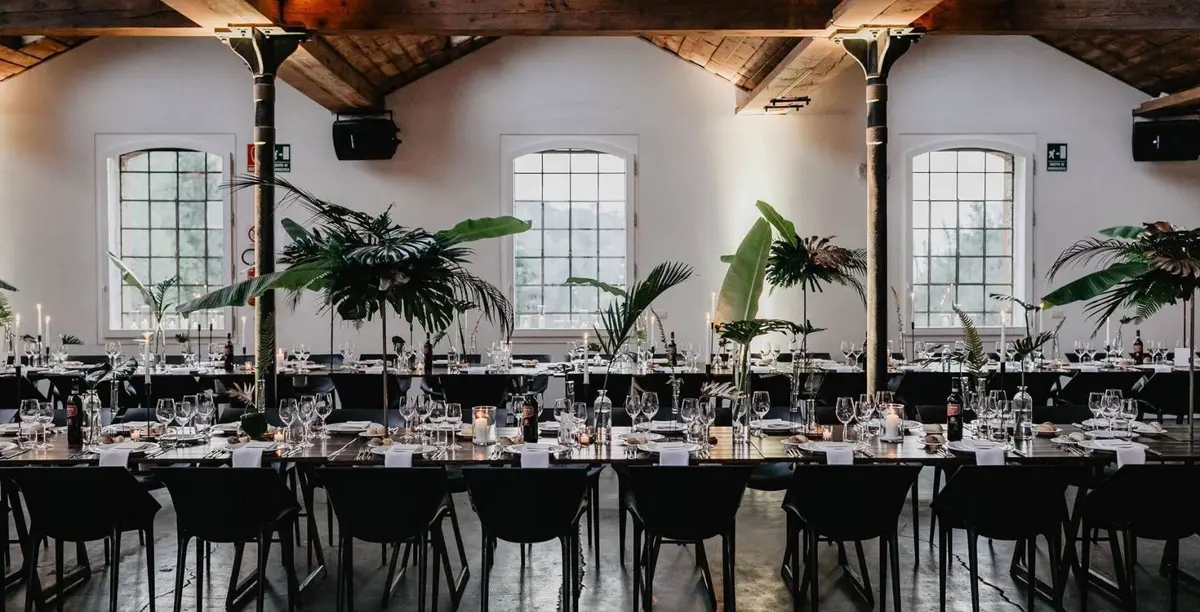 An industrial-chic reception hall: exposed timber beams, black chairs, tall banana-leaf centrepieces
