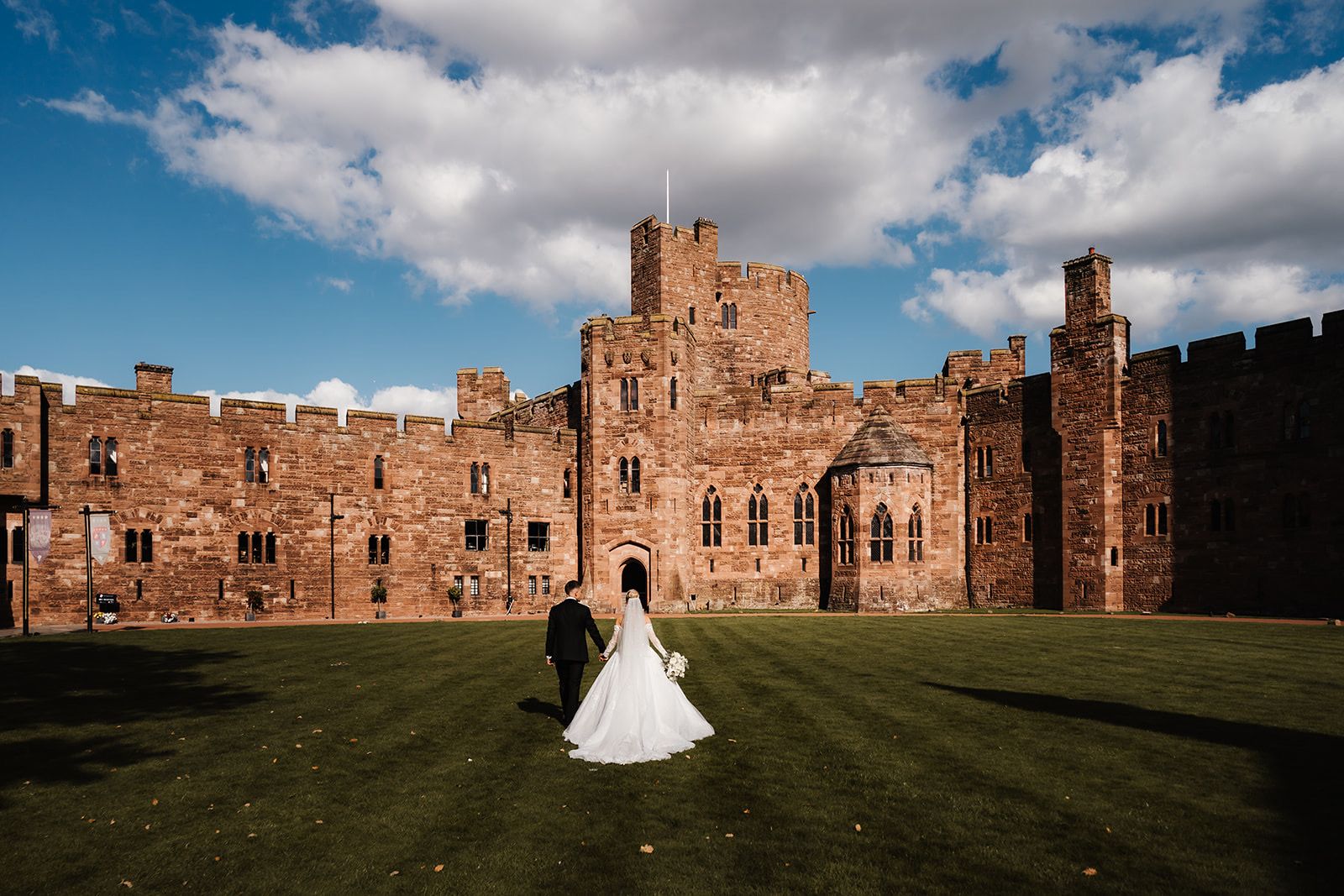 A couple walk across the lawn toward the castle under a bright Cheshire sky