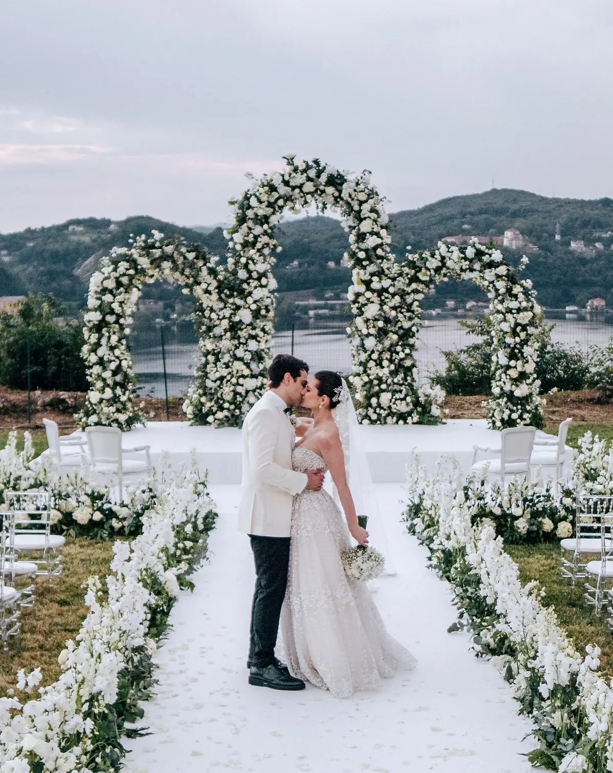 A white double arch above the lake, an aisle of petals, the couple at its head