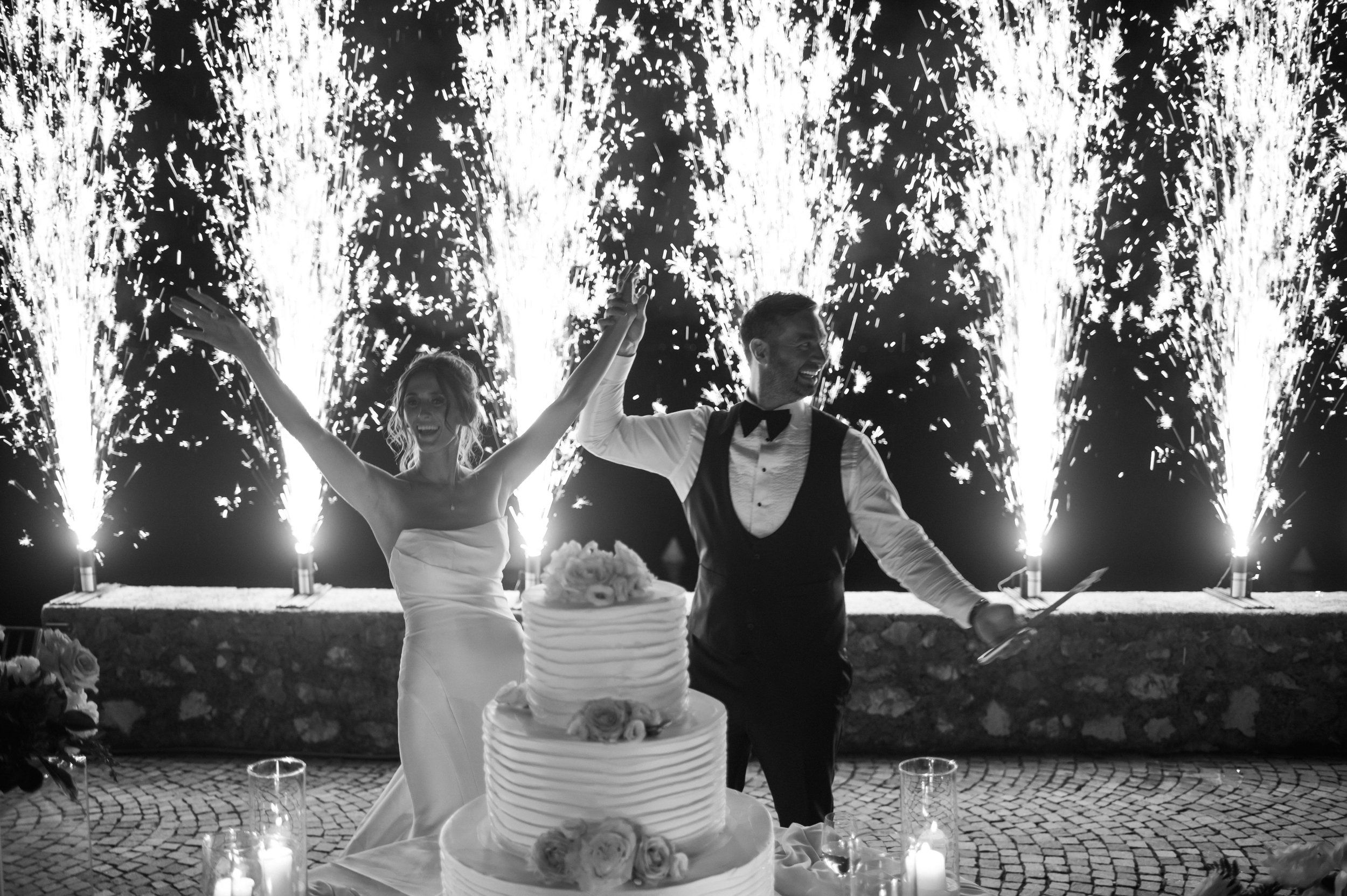 Black and white: a bride and groom with arms raised in front of a three-tier cake as cold-spark fountains erupt behind them