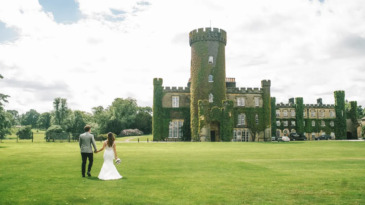 A couple walk hand in hand across the parkland lawn toward the ivy-clad castle and its round tower, Swinton Park, Masham, England