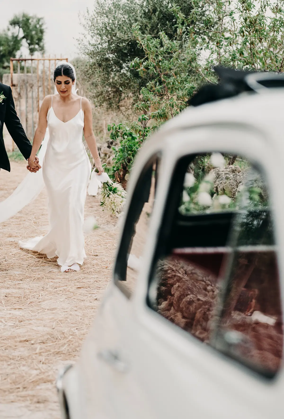 The couple walking a track through olive groves past a white Fiat 500 in Puglia