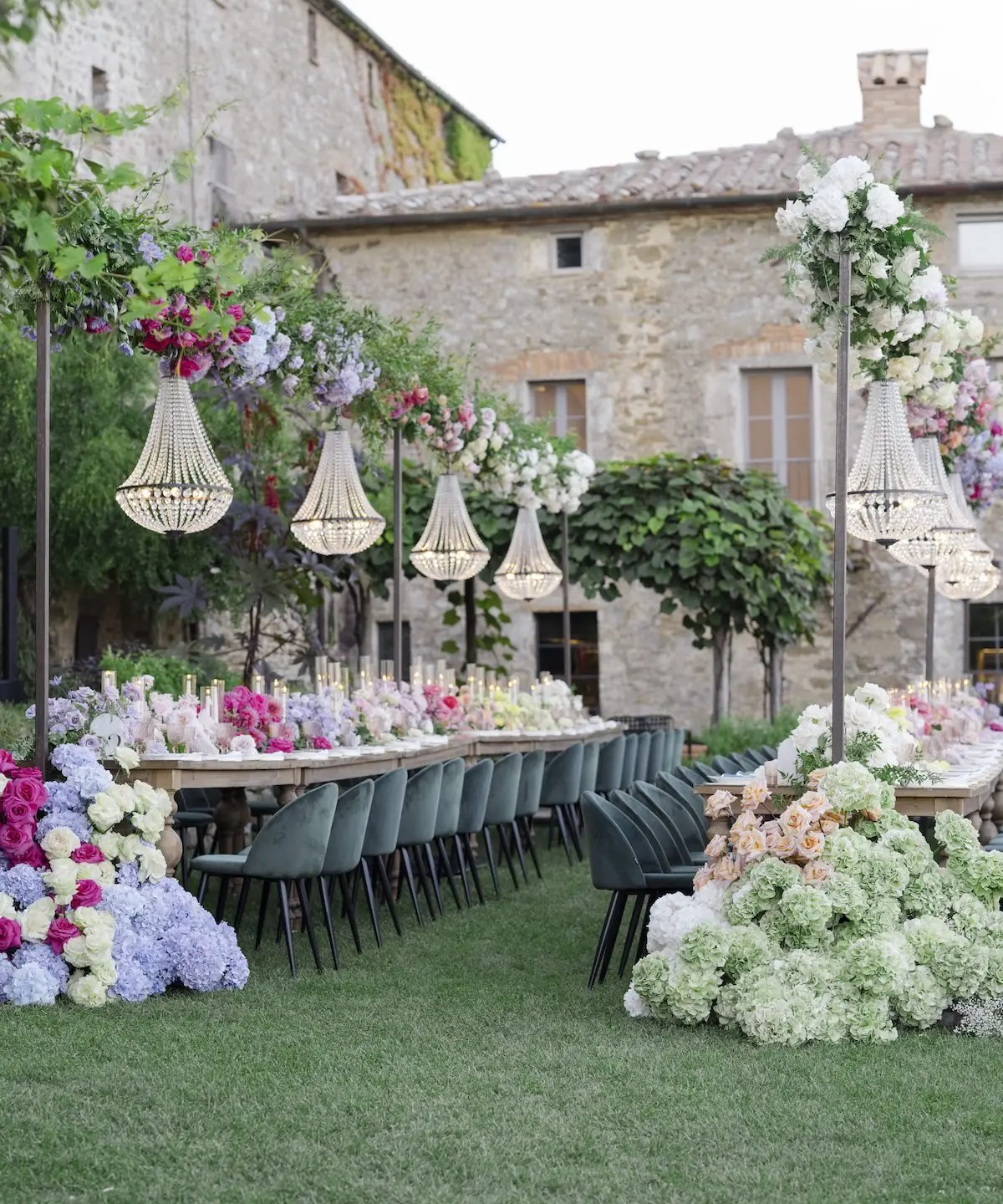 A long table laid on the lawn before a stone borgo, woven pendant lamps overhead