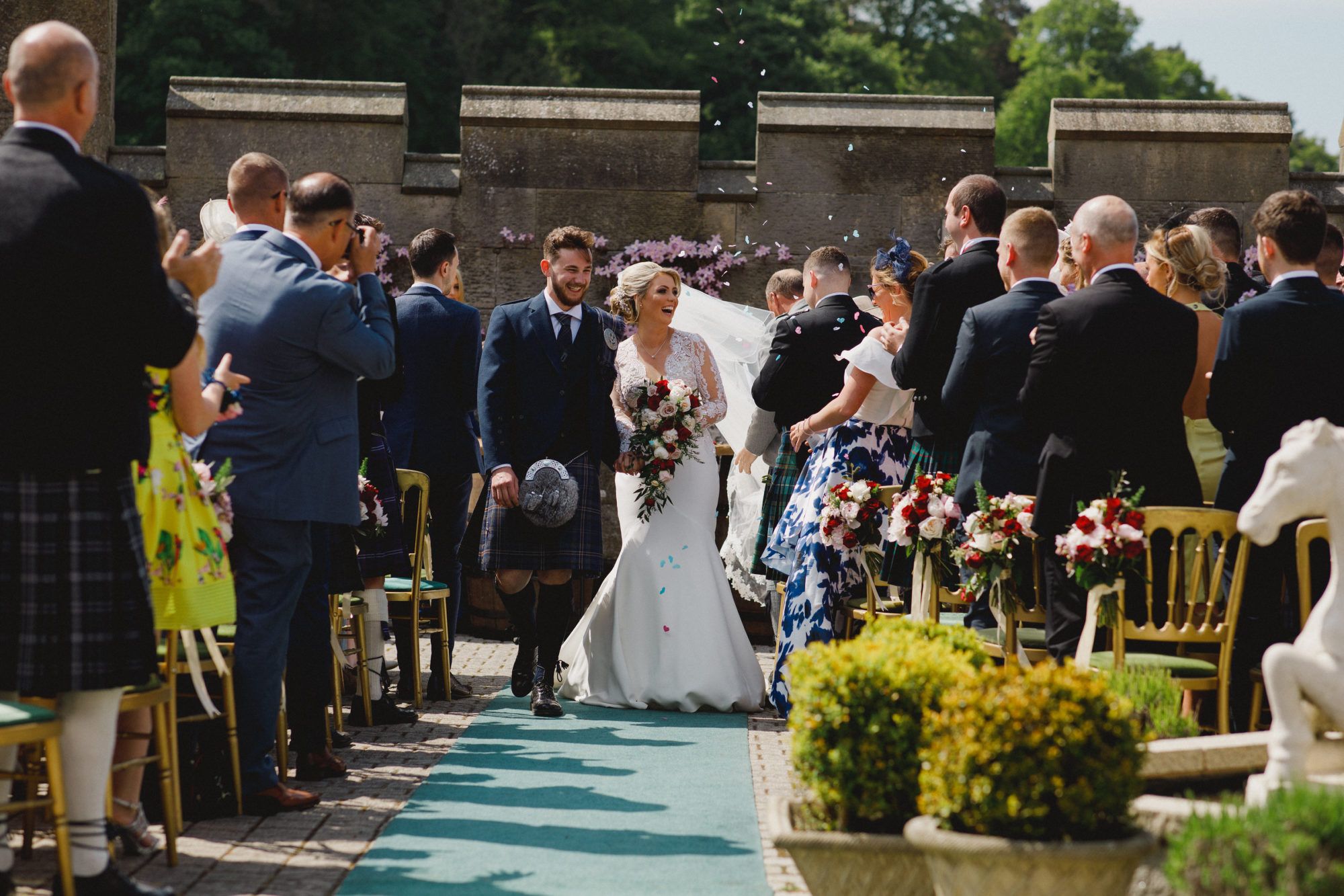 A bride and groom at the front of a courtyard ceremony, a kilted guest among those standing