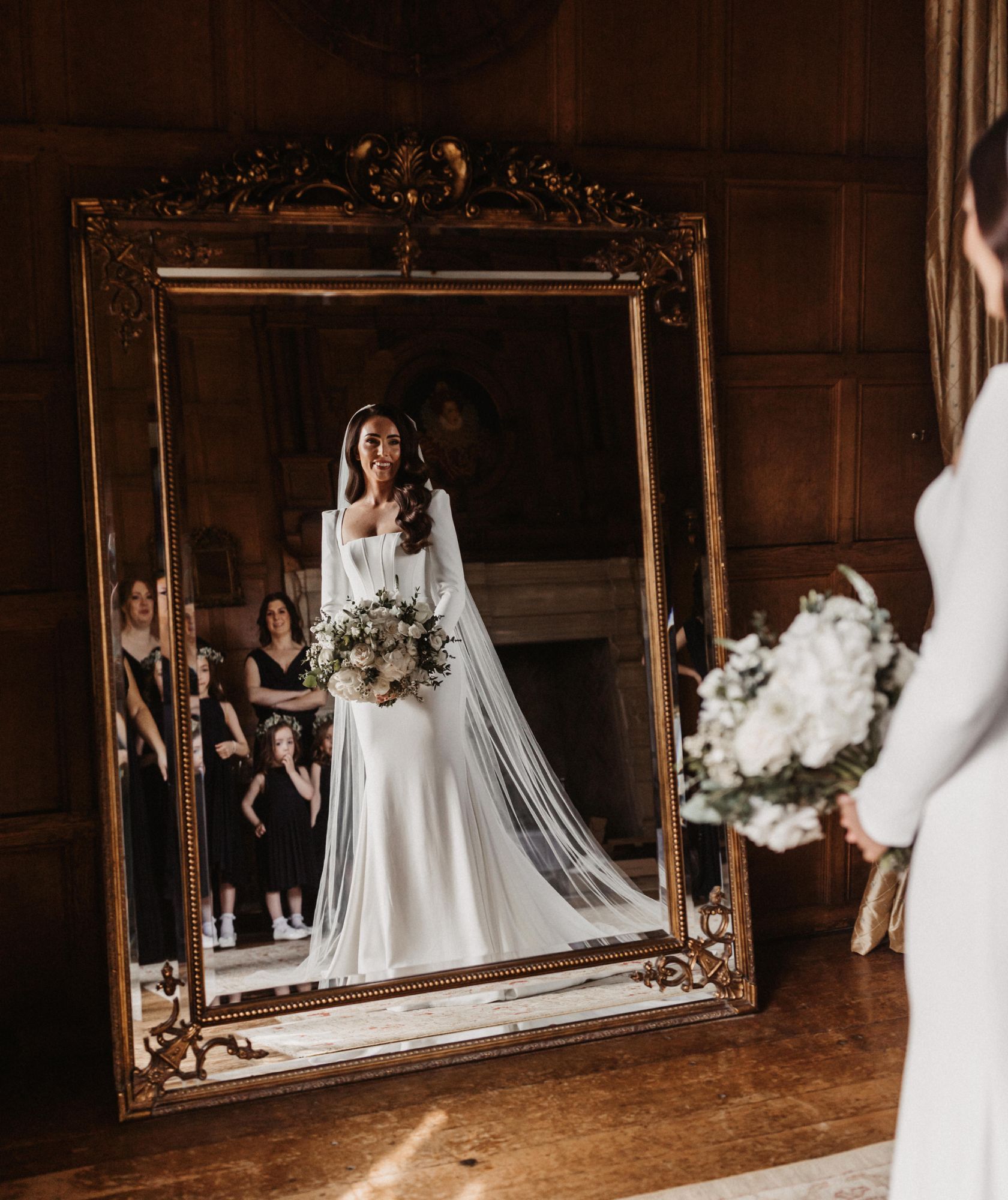 A bride reflected in a large gilt mirror in a panelled room with a stone fireplace