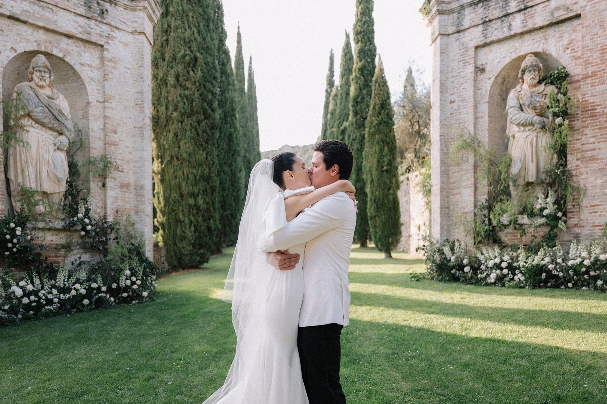 A kiss in a cypress-walled avenue between two brick niches, white roses banked at their feet