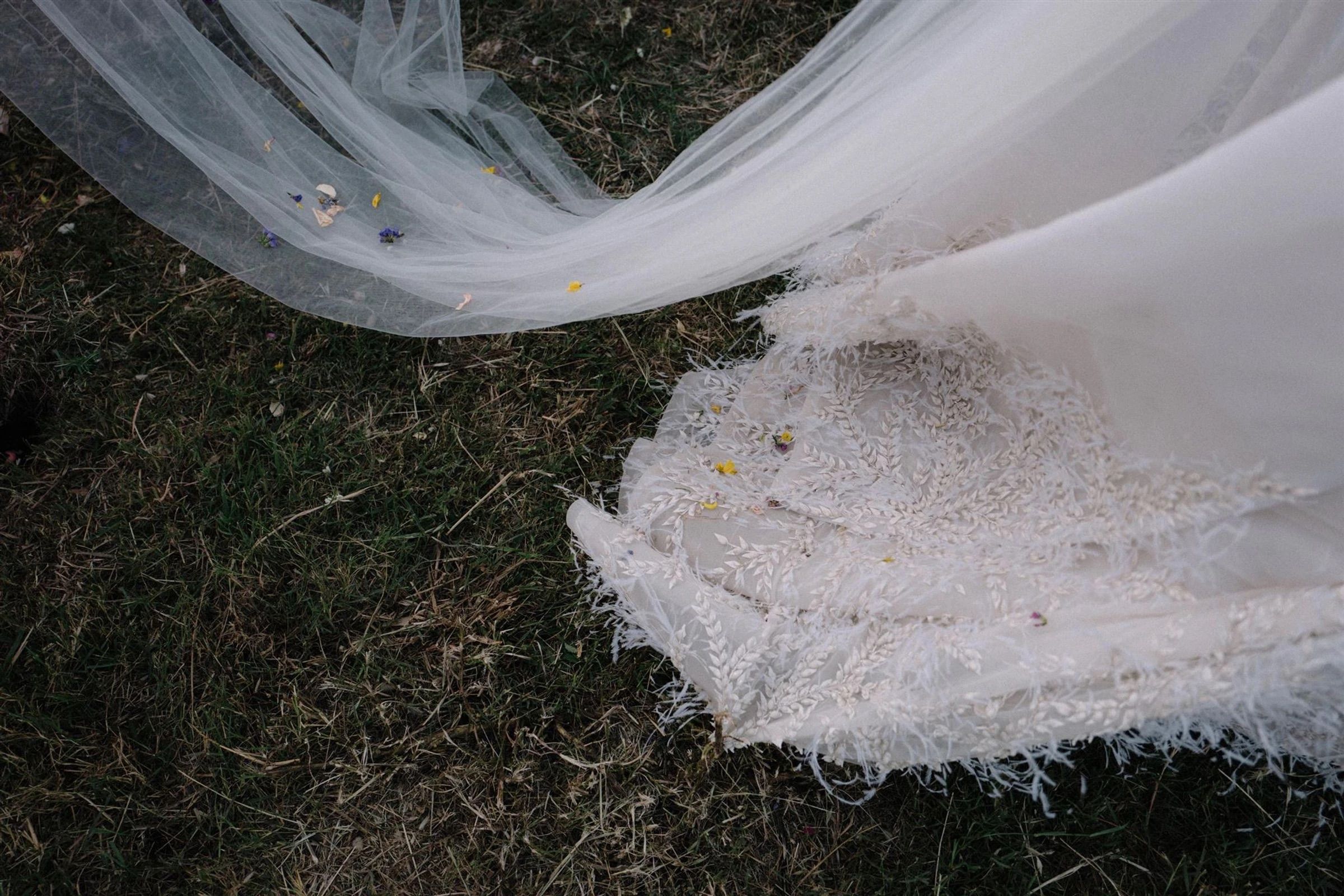 The bride's veil and the lace, feather-trimmed hem of her gown lie spread across the grass