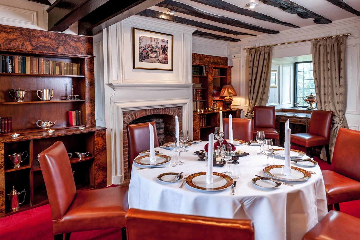 A panelled dining room lined with books, a round table laid for dinner by the fire