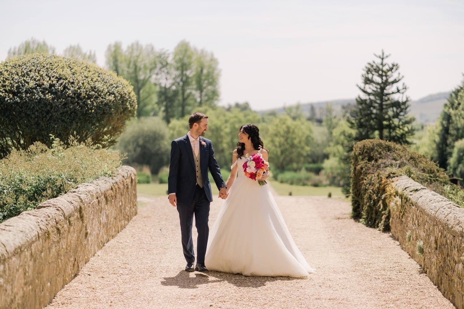 A bride and groom on a stone causeway across the moat, the hills rising behind