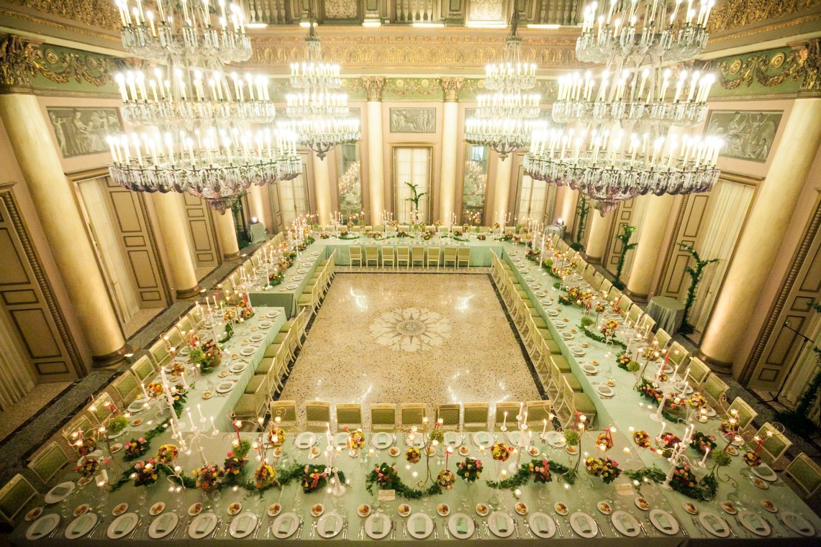 A hollow rectangle of table under the chandeliers of a gilded ballroom