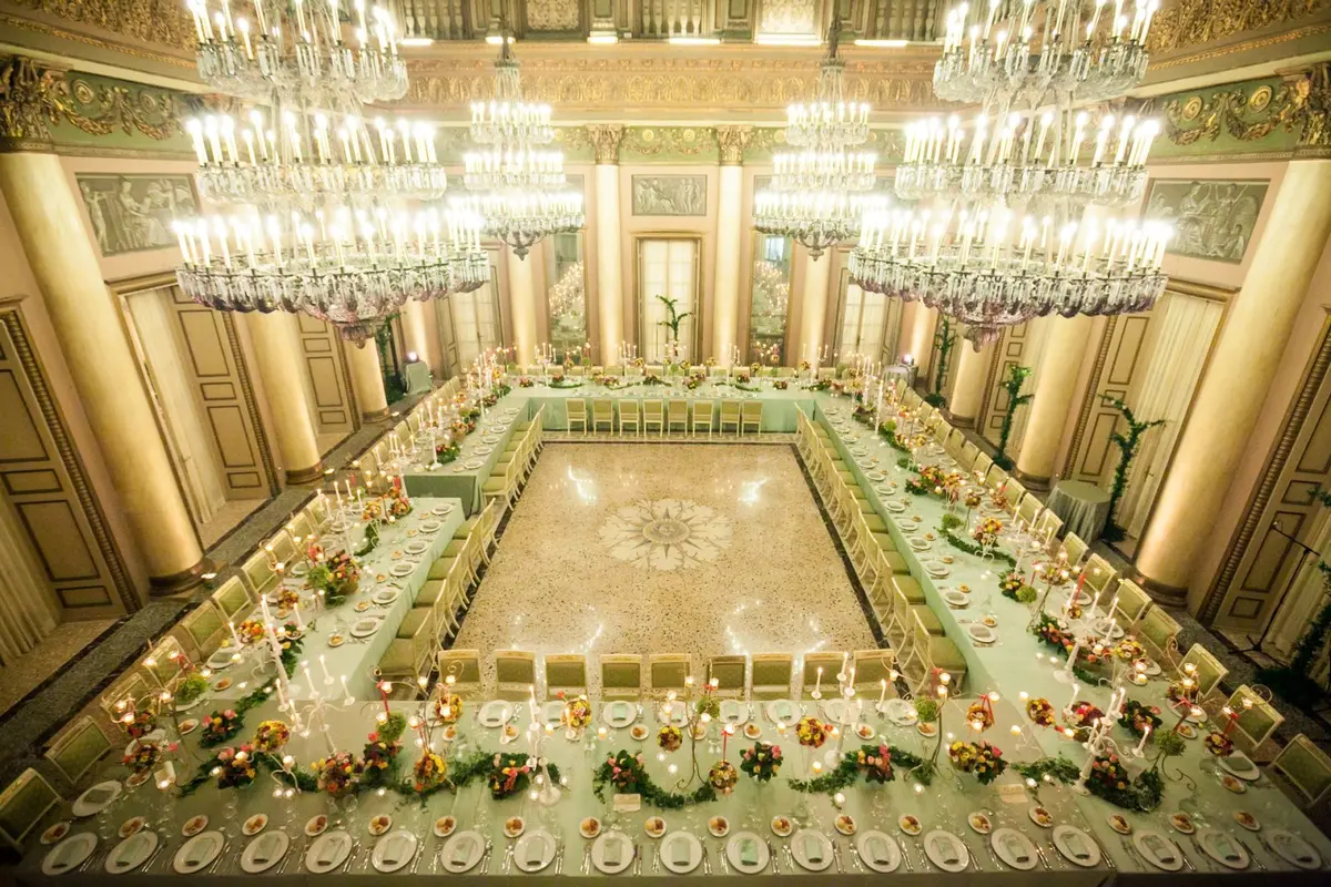 A hollow rectangle of table under the chandeliers of a gilded ballroom