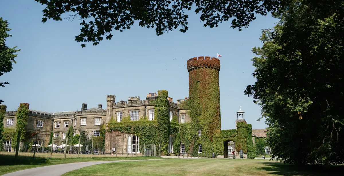 The castle exterior, the ivy-clad round tower rising above the castellated wings on a summer day, Swinton Park, Masham, England